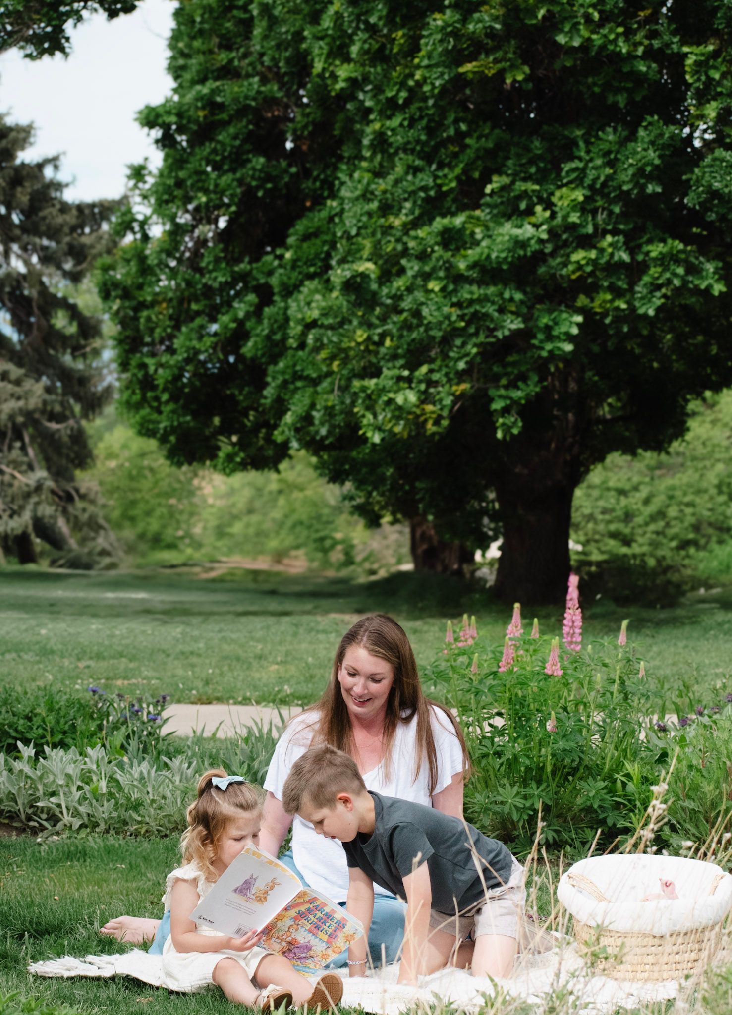 A mother sits on a blanket in a lush green park, reading a book with her two young children on a sunny day.