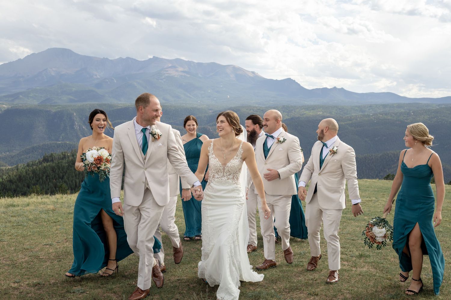 Wedding party walks outdoors with stunning mountain backdrop, bride in white gown, bridesmaids in teal dresses, groomsmen in tan suits.