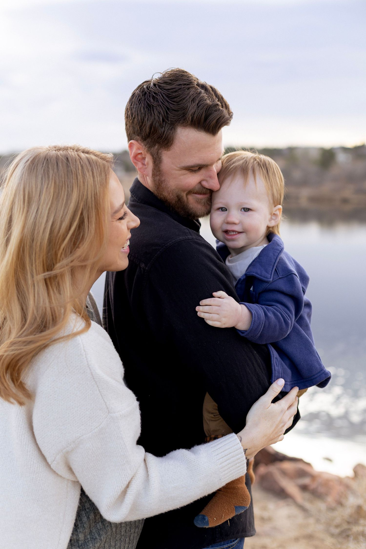 A happy young family with a toddler boy poses near a lake outdoors in cool weather.