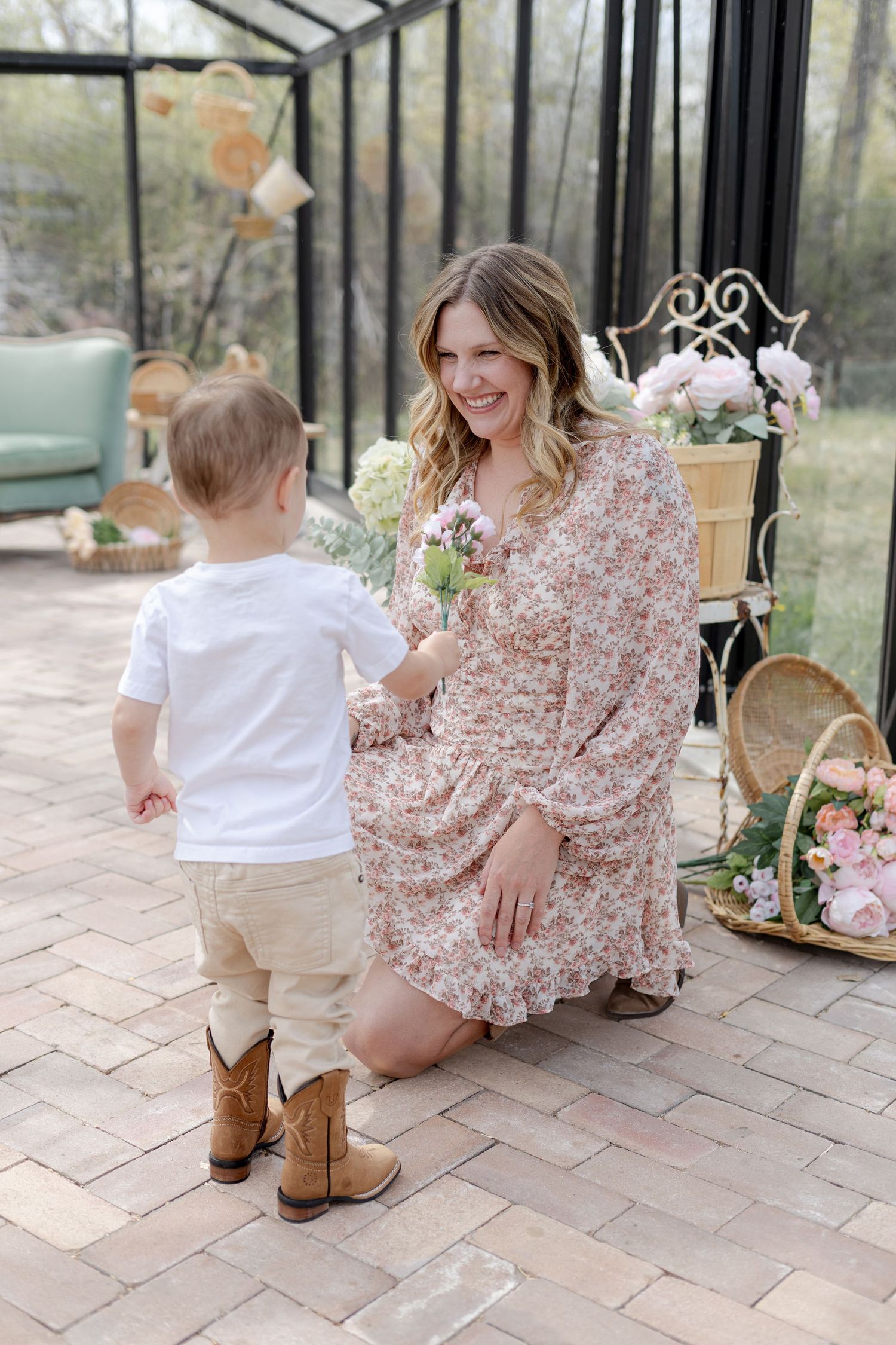 A smiling woman in a floral dress kneels as a toddler in cowboy boots offers her a flower in a greenhouse setting.