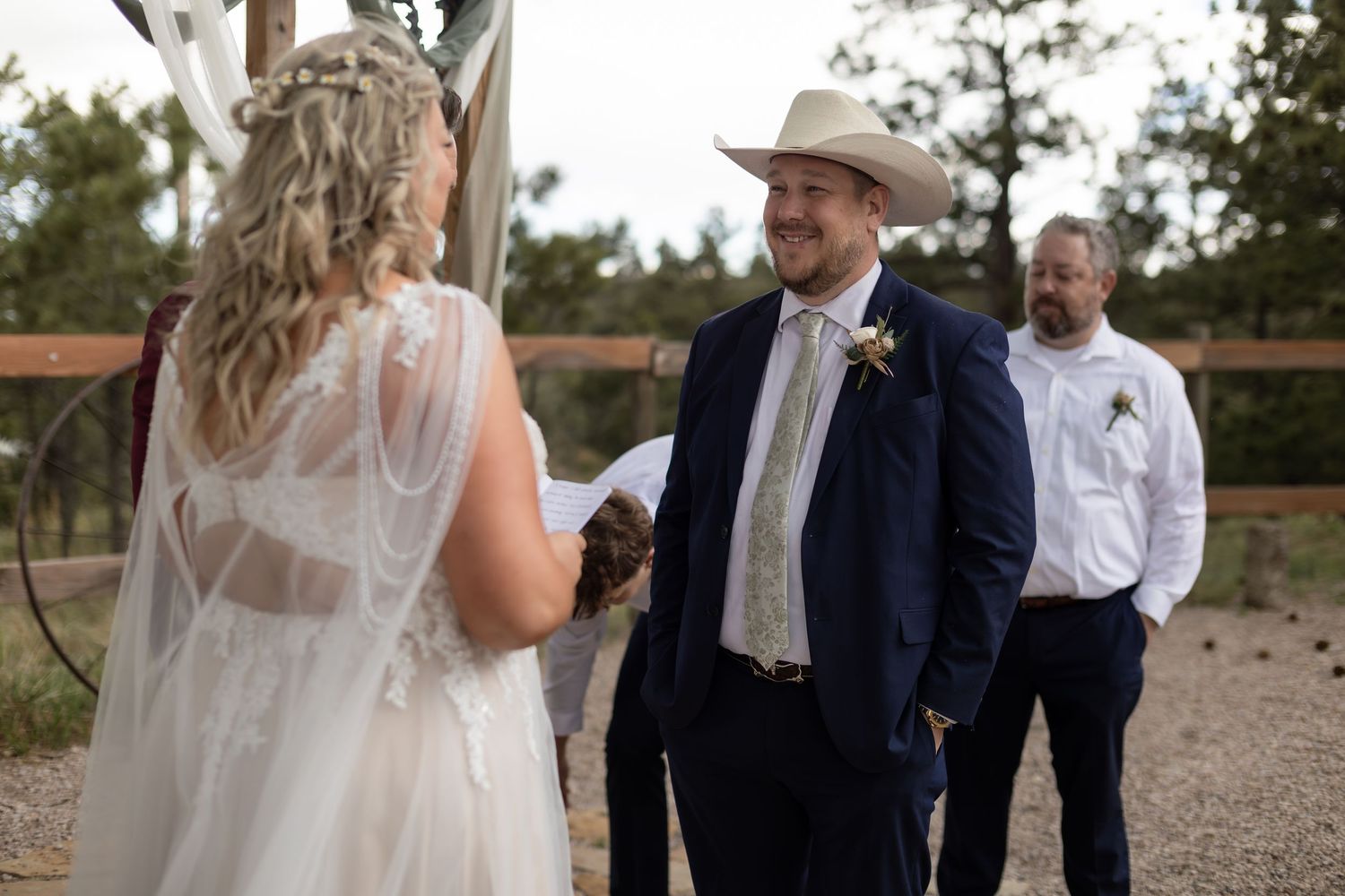 Groom in navy suit and cowboy hat smiles at bride during outdoor rustic wedding ceremony.