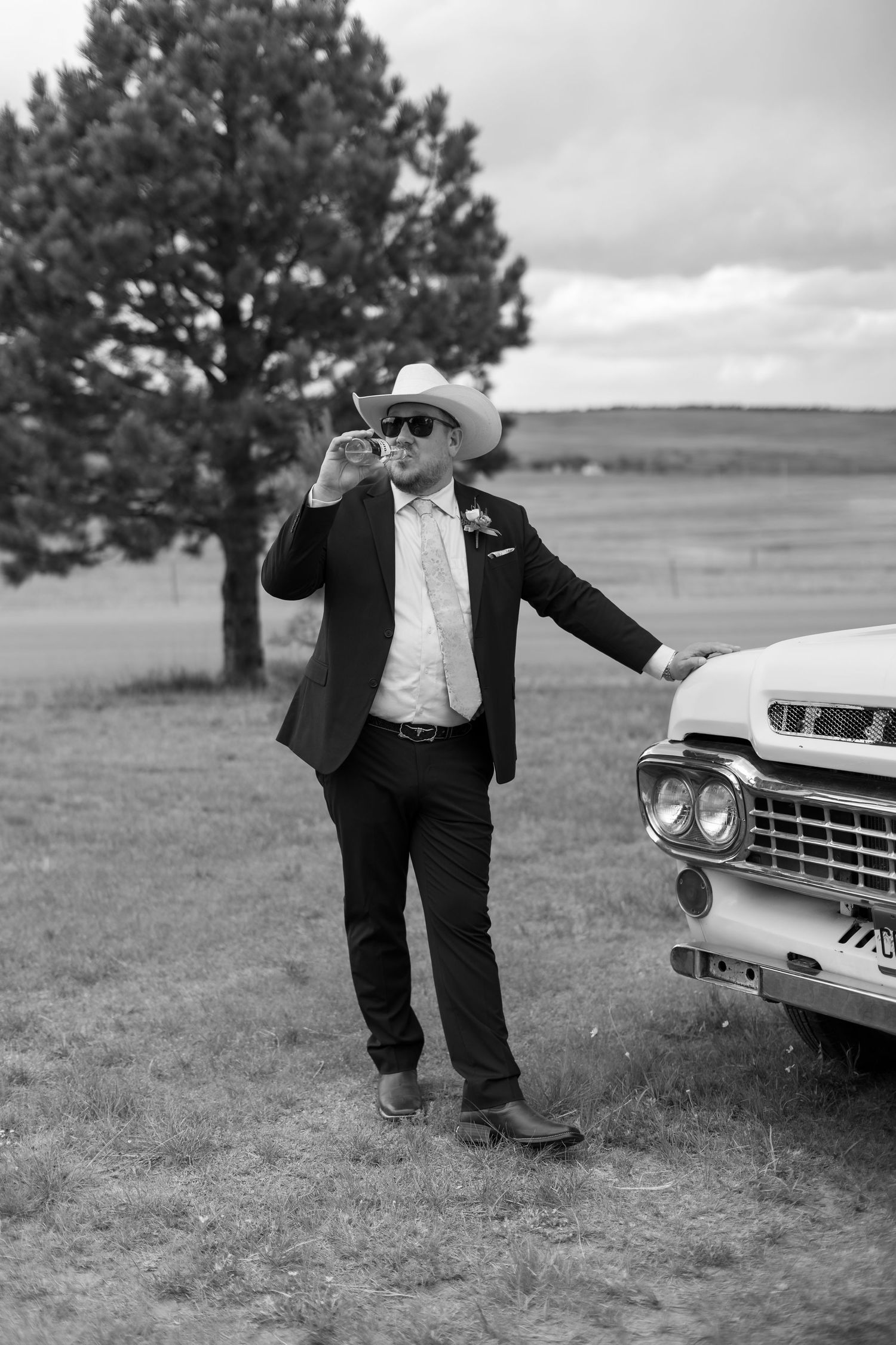 Groom in cowboy hat and suit smoking cigar leaning on vintage classic car in black and white photo.