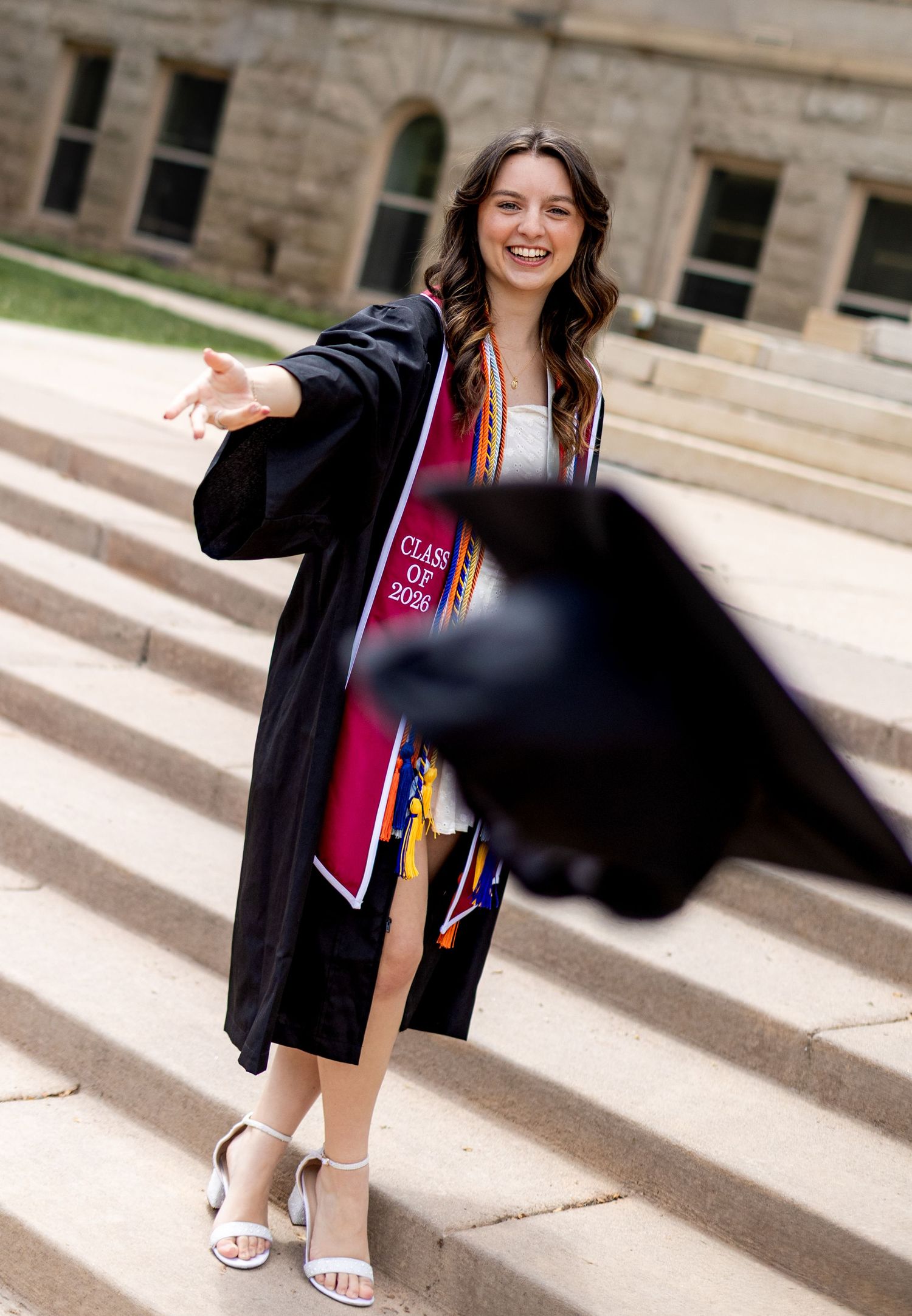 A smiling female graduate in black gown and red Class of 2025 sash tosses her cap on stone steps.