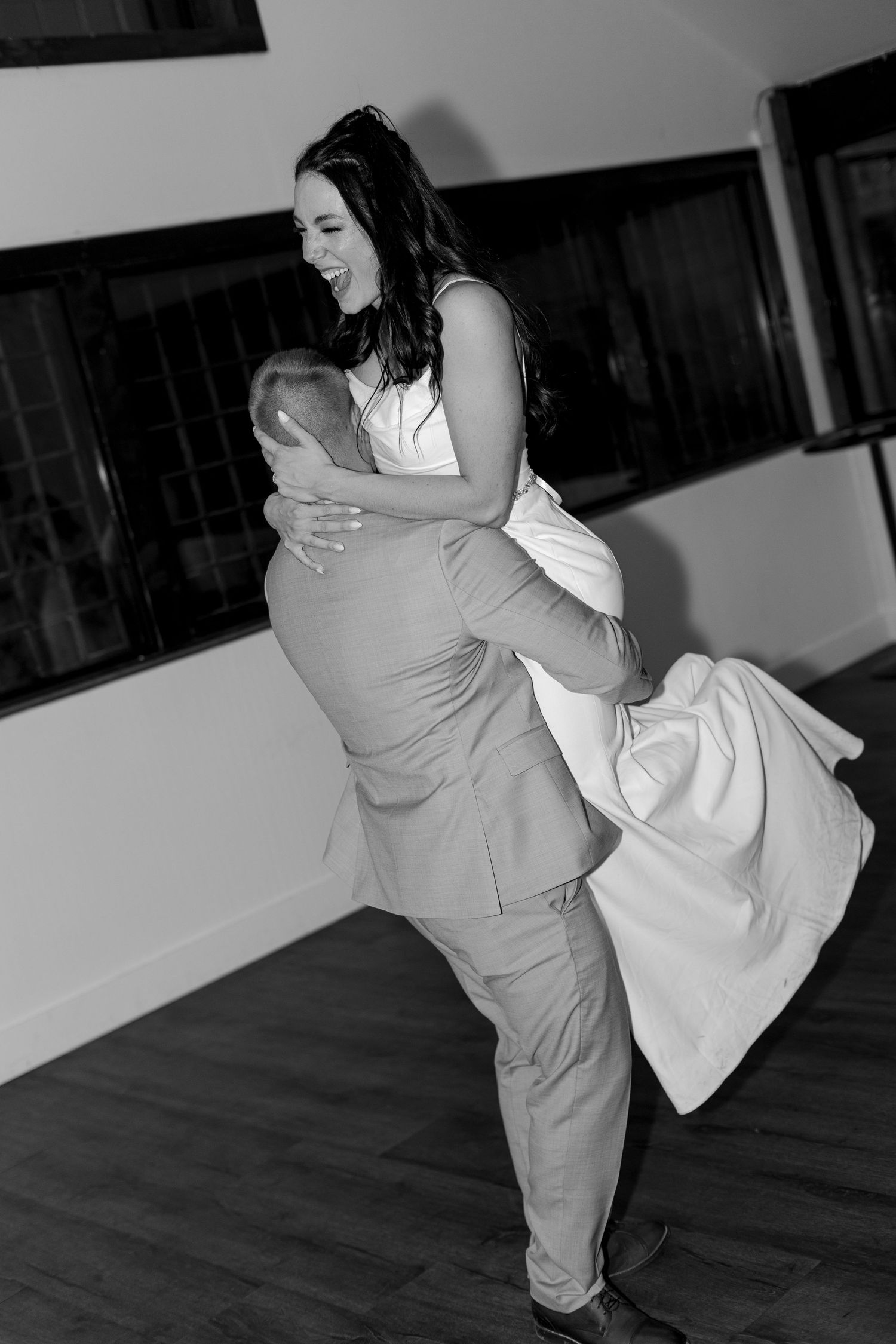 Black and white photo of groom lifting bride joyfully during first dance at elegant wedding reception.