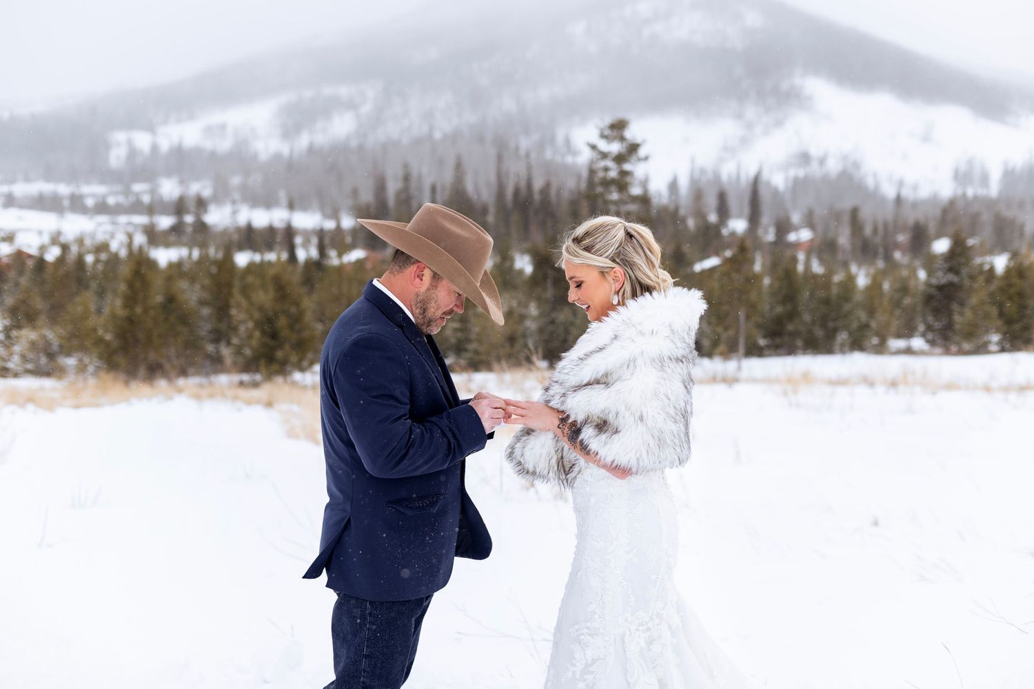 A couple shares a romantic moment in a snowy winter landscape with snow-covered mountains in the background.