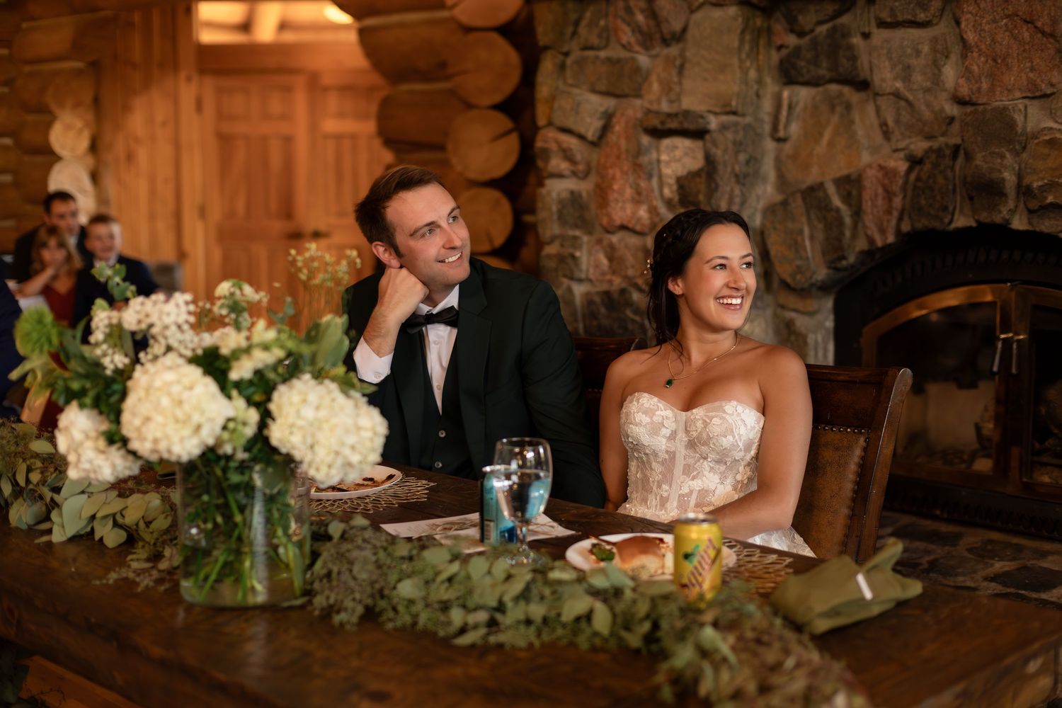 Bride and groom smiling at sweetheart table with white floral centerpiece in rustic log cabin wedding venue.