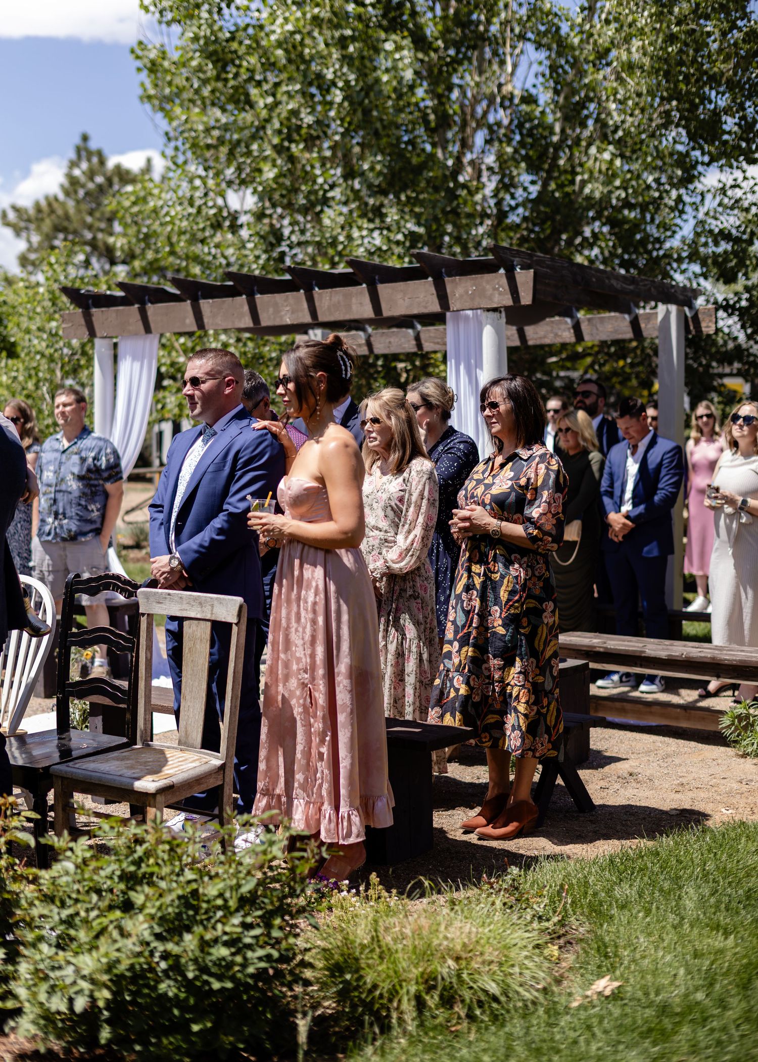 Wedding guests standing and watching an outdoor ceremony near a wooden pergola draped with white fabric.