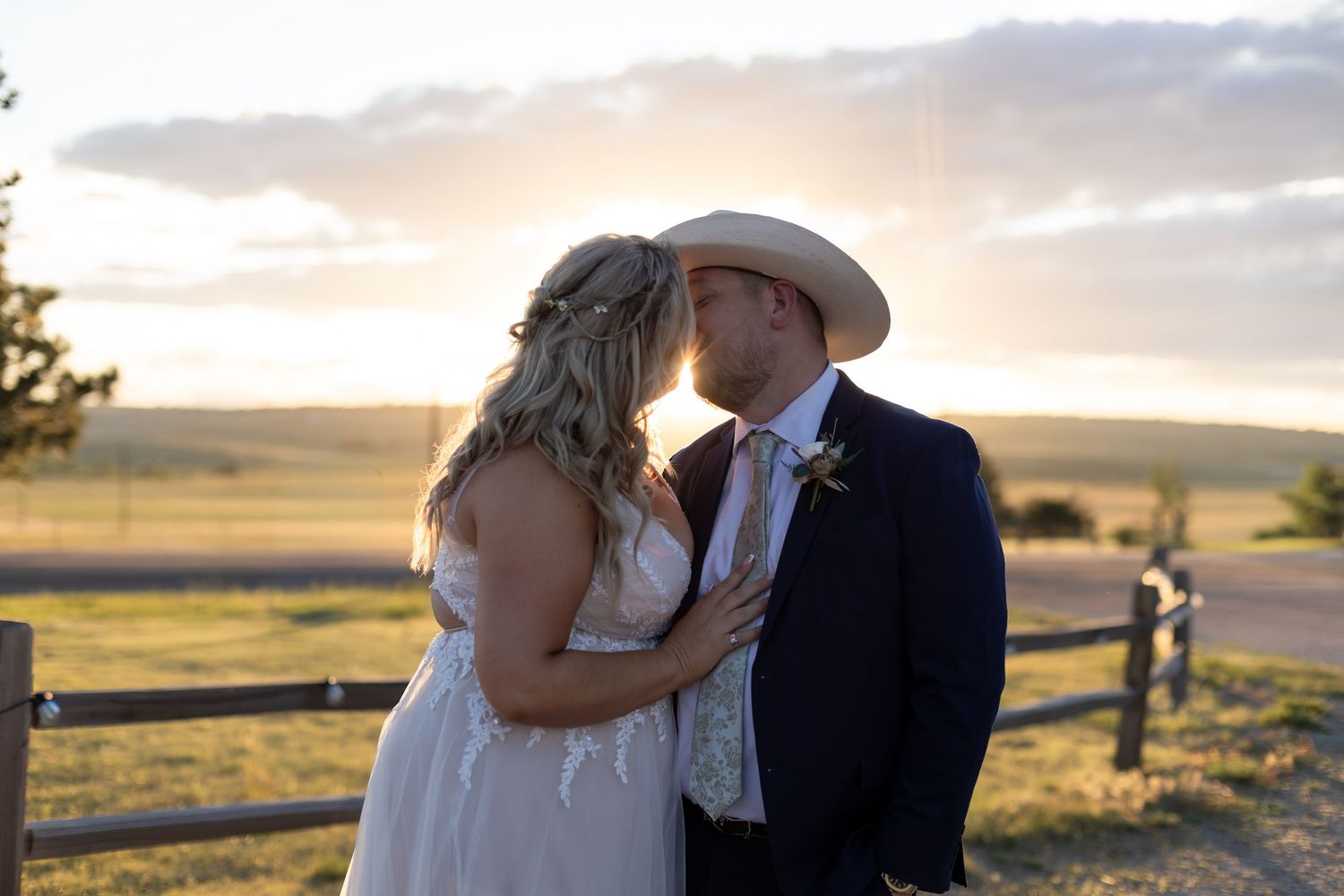 Bride and groom share a kiss at golden hour near a rustic fence in an open field during their outdoor country wedding.
