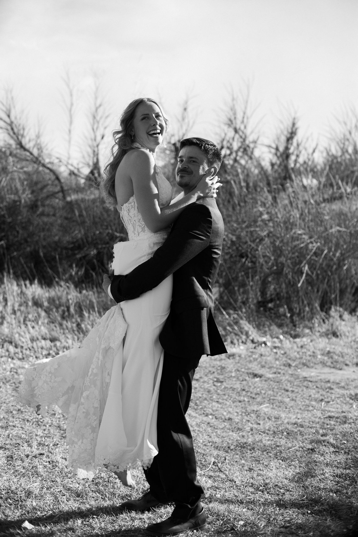Black and white photo of joyful groom lifting laughing bride outdoors in natural setting.