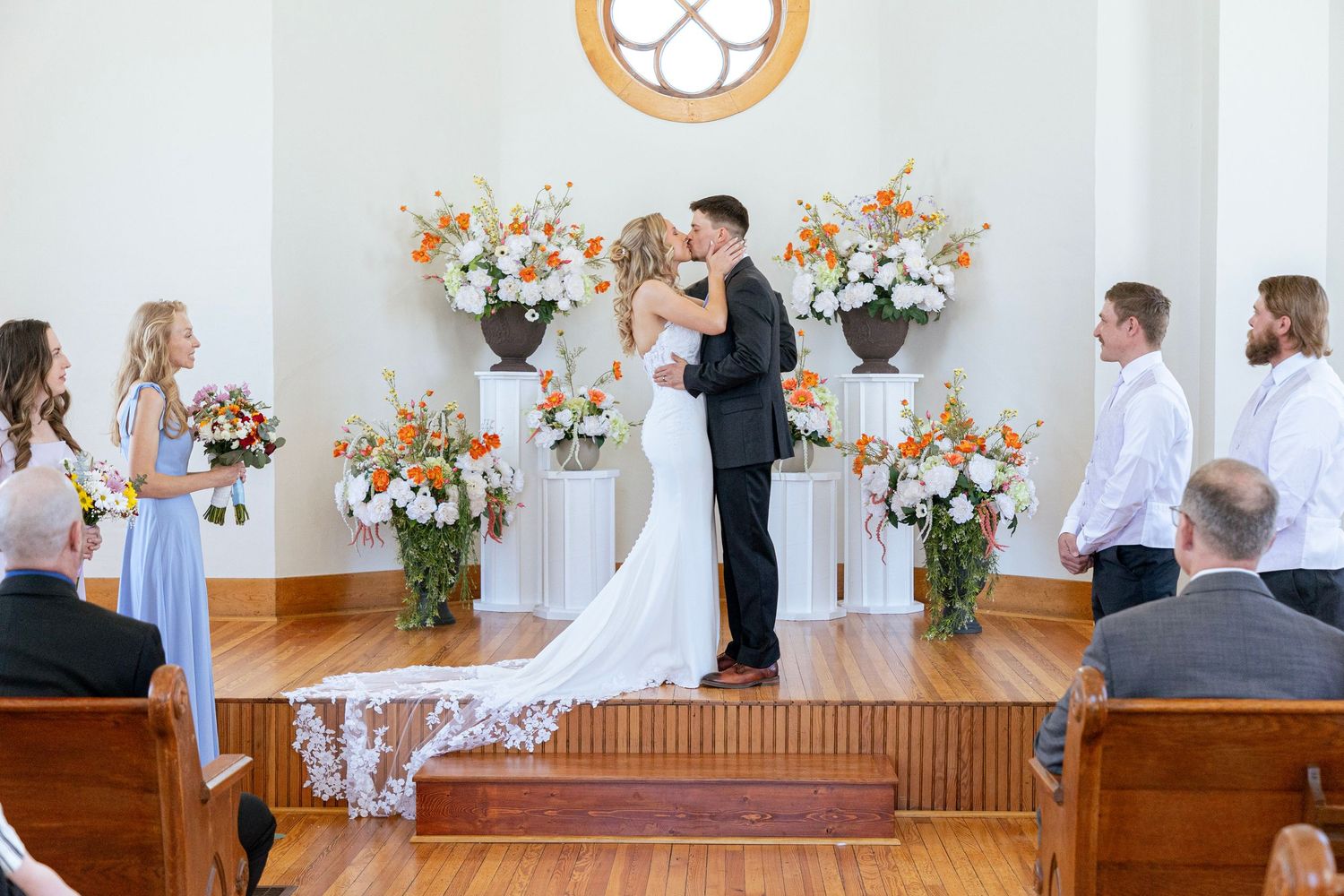 Bride and groom share first kiss at altar adorned with orange and white floral arrangements in a bright white chapel.