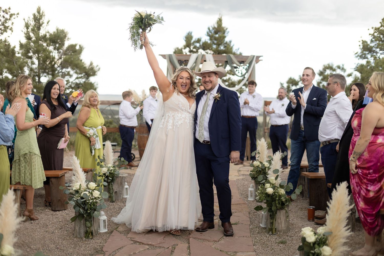Joyful bride and groom in cowboy hat walk back down aisle as guests celebrate at outdoor rustic wedding.