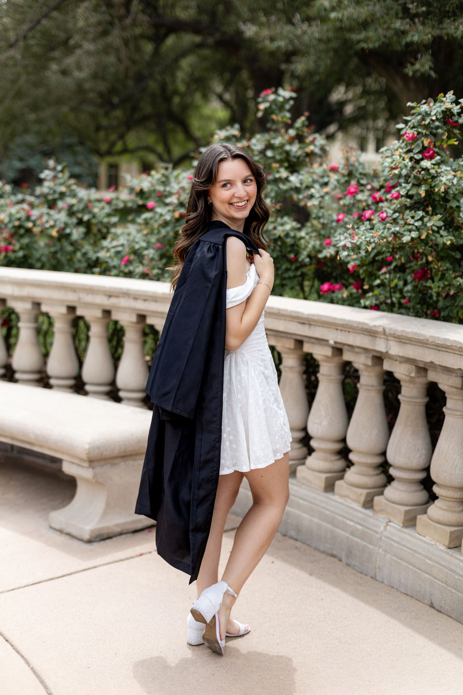 Smiling young woman in white dress holding graduation gown by stone balustrade with blooming flowers.