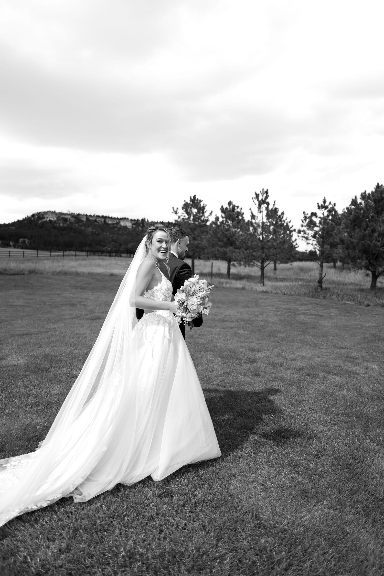 Black and white photo of smiling bride in flowing gown holding bouquet with groom embracing her in a field.