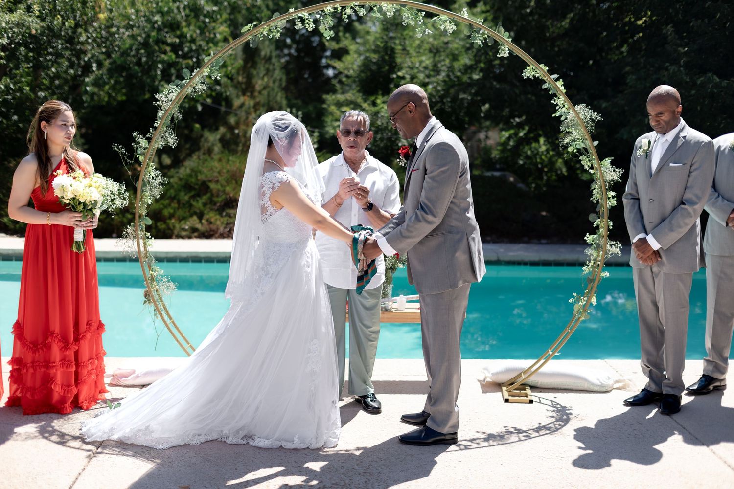 Outdoor wedding ceremony by a pool with bride in white gown, groom in brown suit, officiant, bridesmaid, and groomsman.