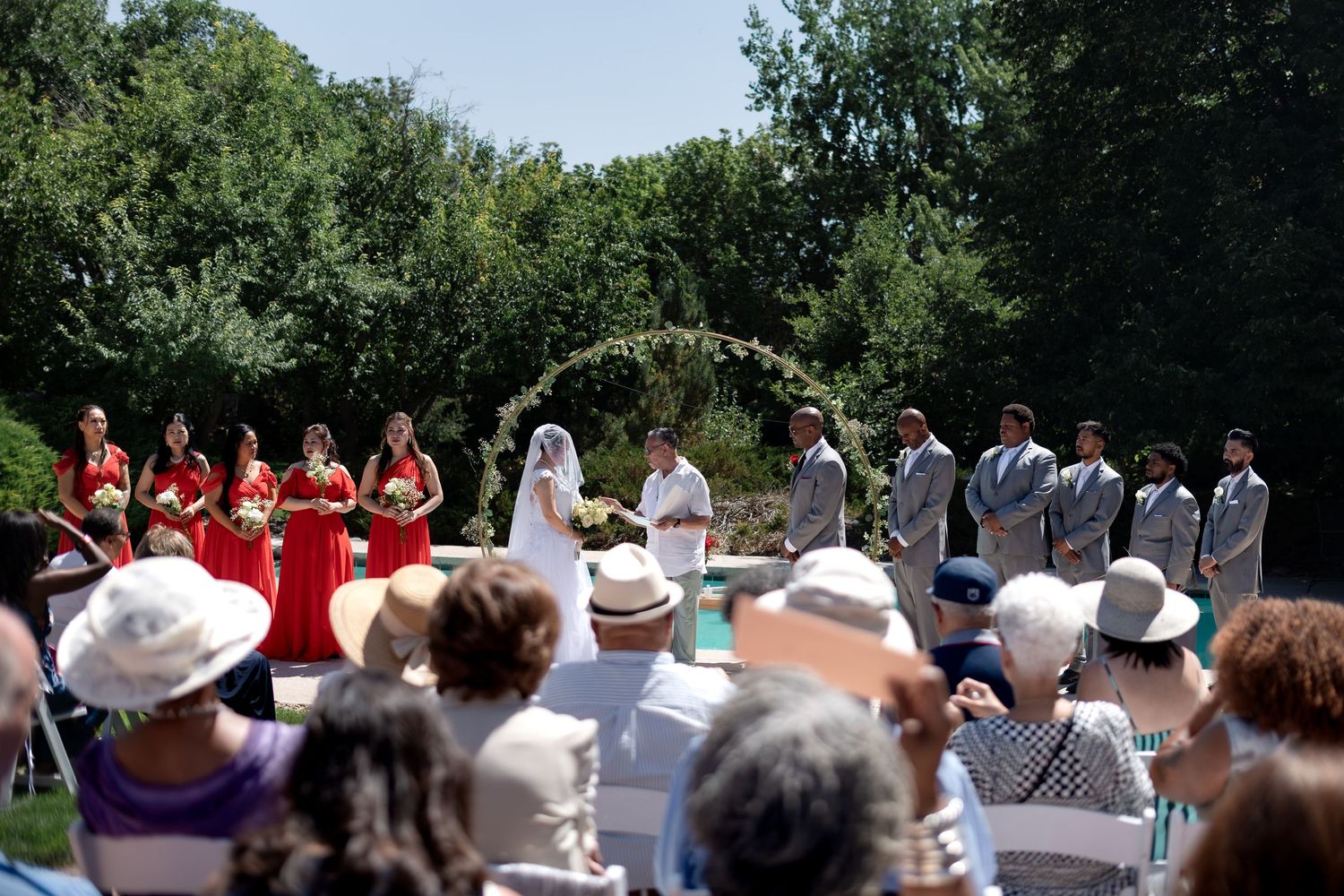 Outdoor wedding ceremony with red bridesmaids, groomsmen, and couple at floral arch, surrounded by lush greenery.