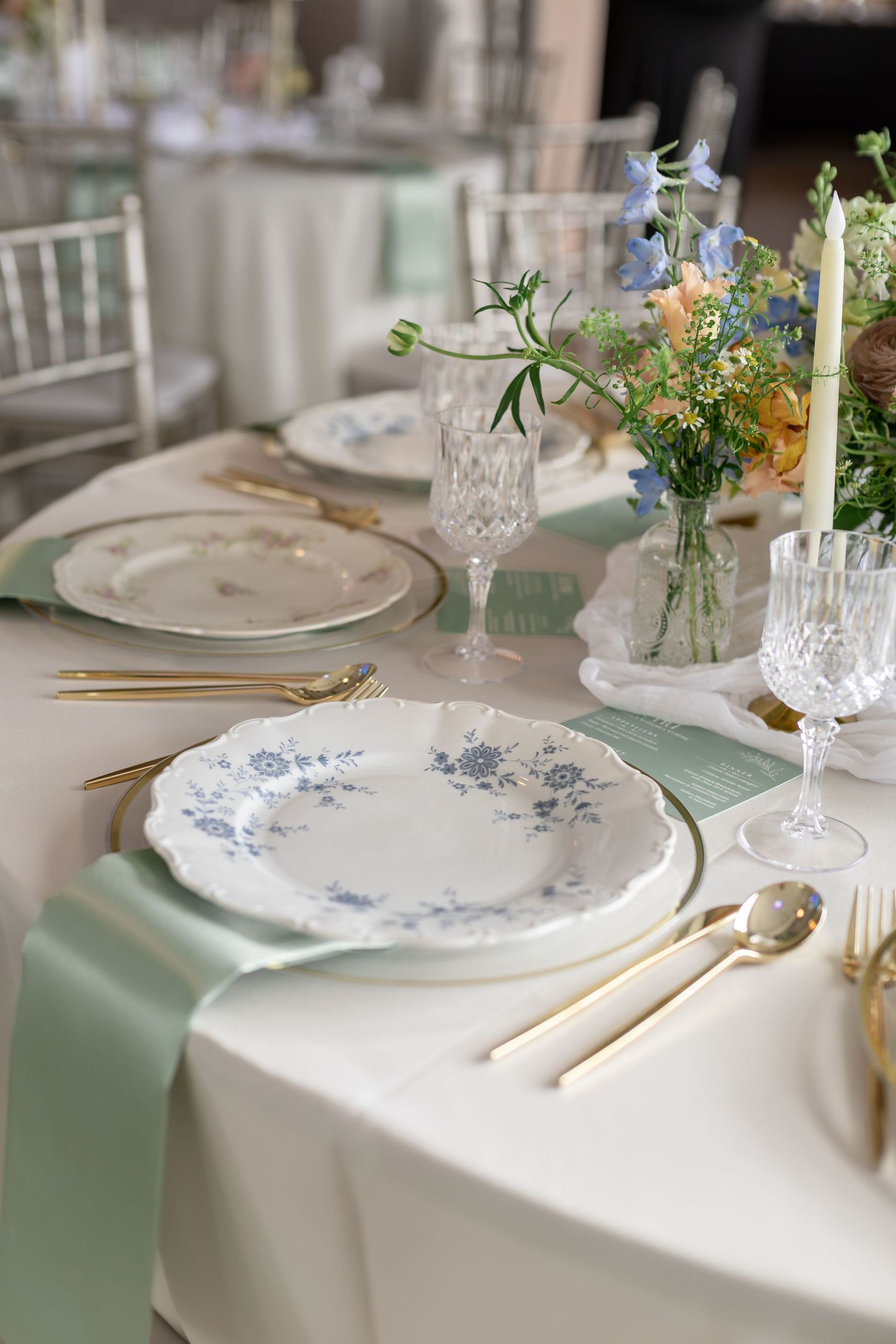 Elegant wedding table setting with blue floral plates, gold cutlery, crystal glasses, and pastel floral centerpieces on white linen.