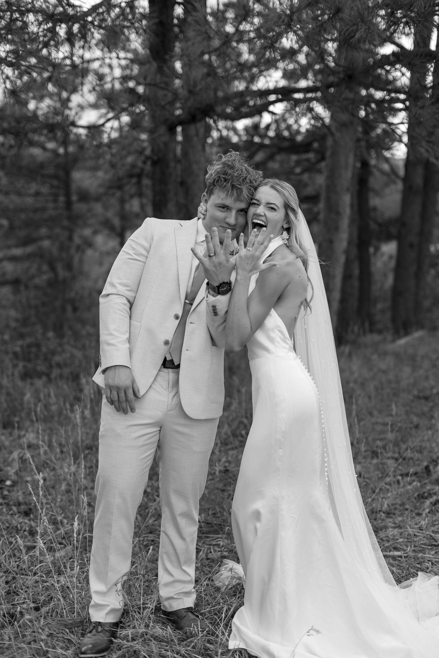 Joyful bride and groom in forest setting showing wedding rings, black and white photo.