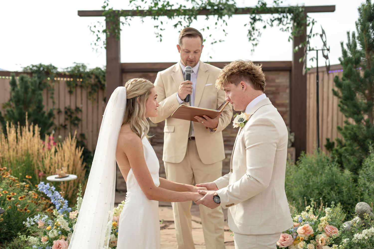 Outdoor wedding ceremony with bride and groom holding hands before officiant, surrounded by colorful floral arrangements.