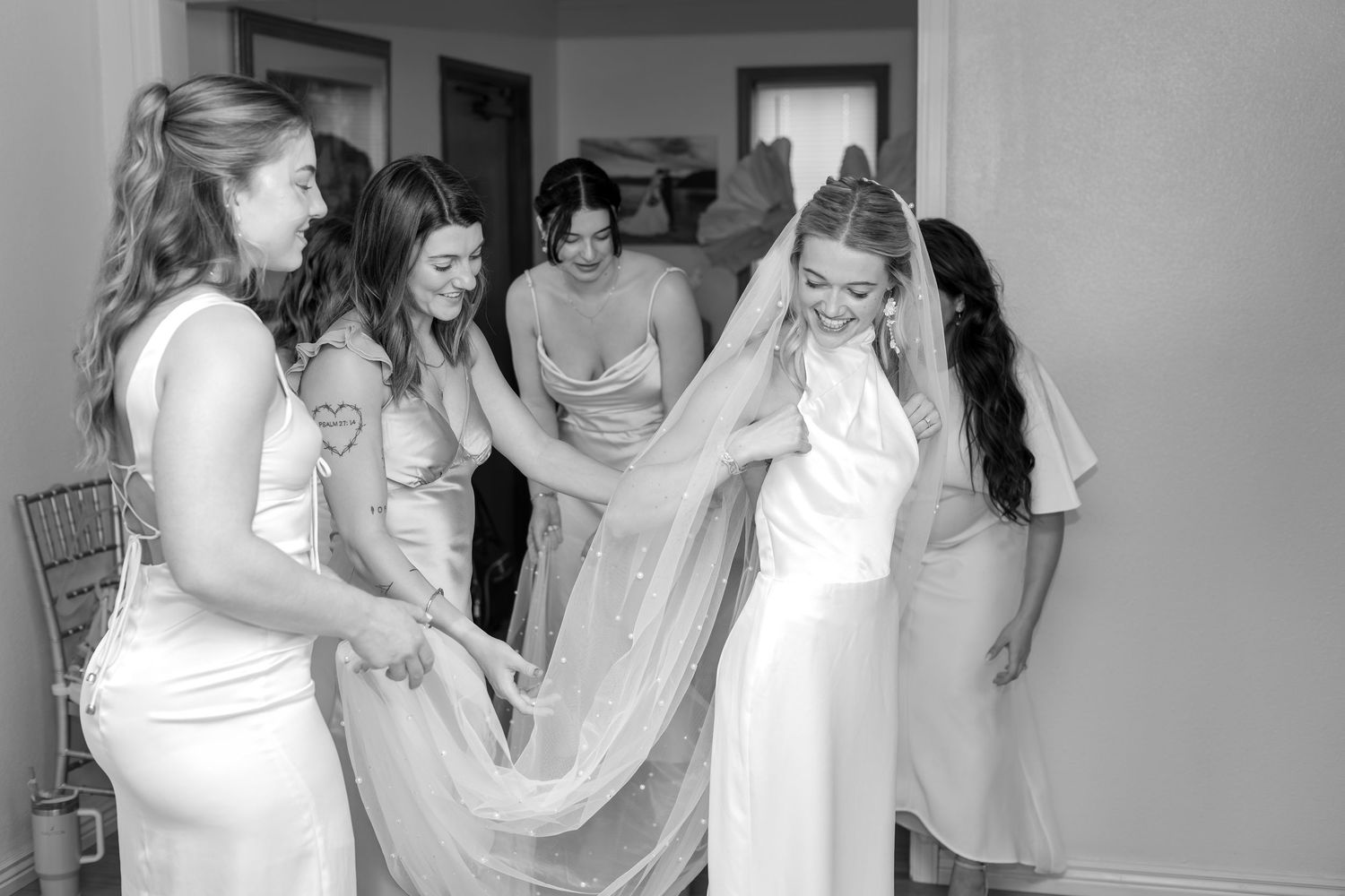 Black and white photo of bride and bridesmaids adjusting wedding veil indoors before ceremony.