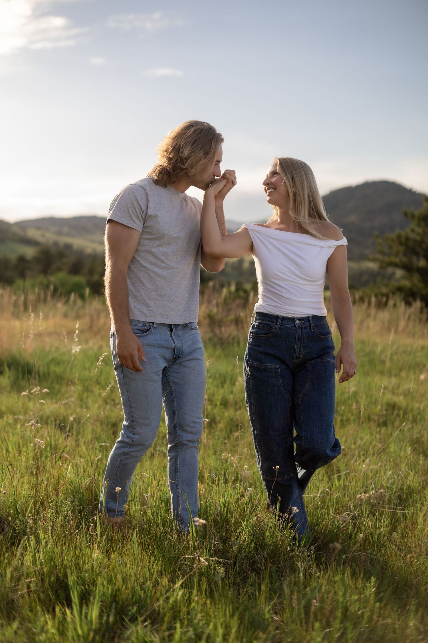 Couple walking hand in hand through green meadow, man kissing woman's hand, rolling hills and blue sky backdrop.