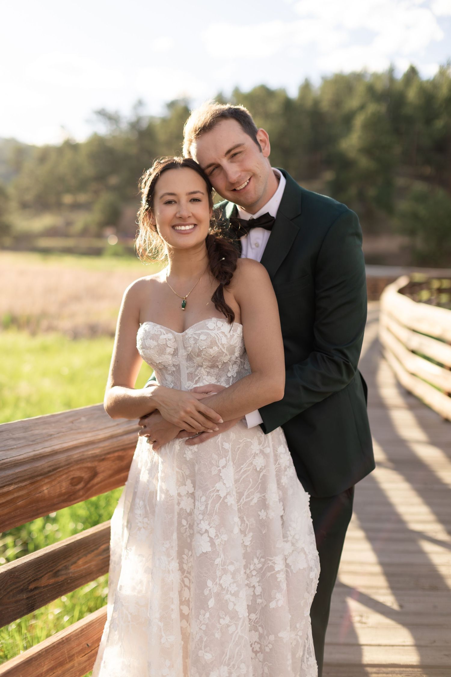 Smiling bride and groom in green tuxedo embrace on wooden bridge at golden hour outdoor wedding.