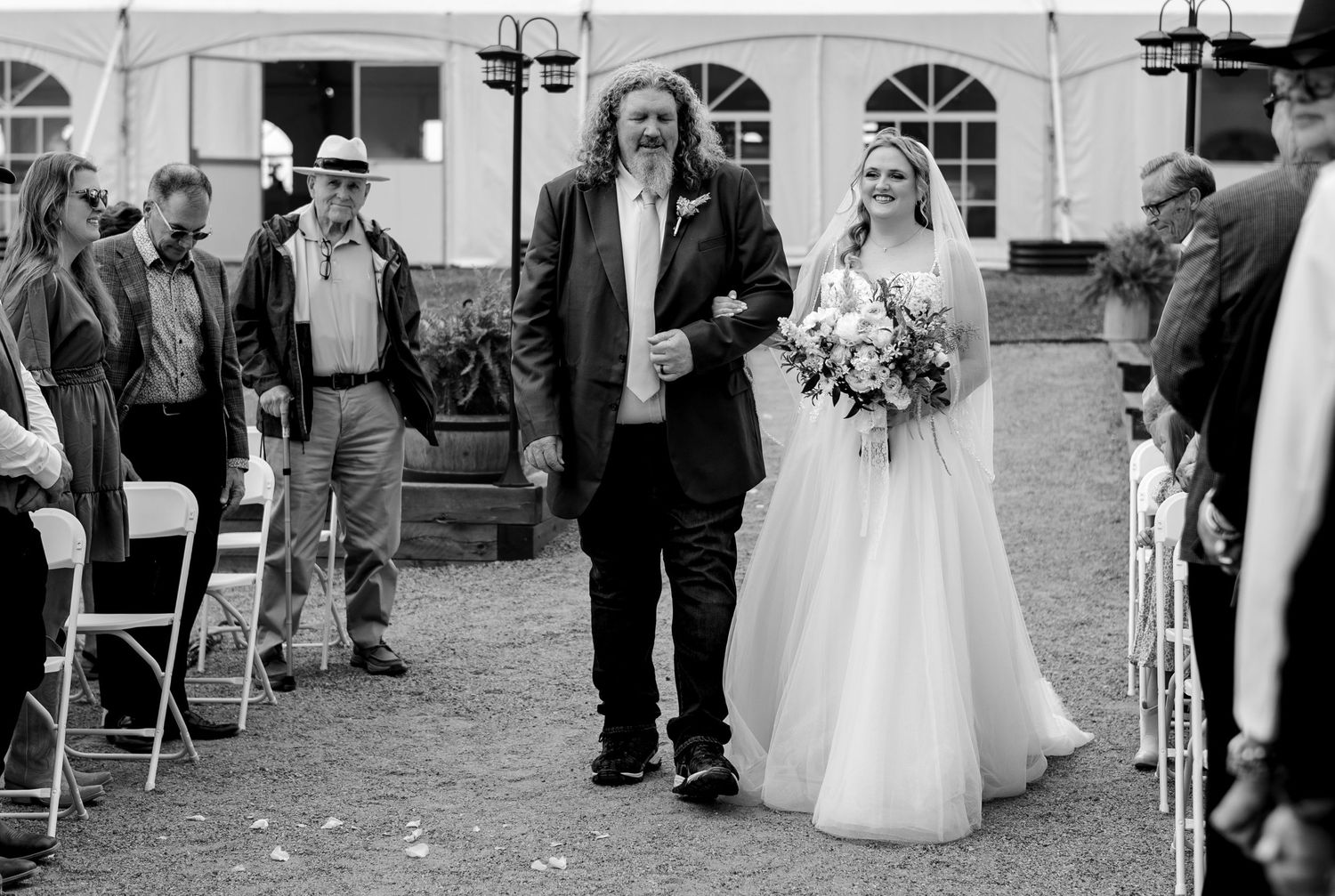 Father walks bride down the aisle at outdoor wedding ceremony, bride holds bouquet in black and white photo.