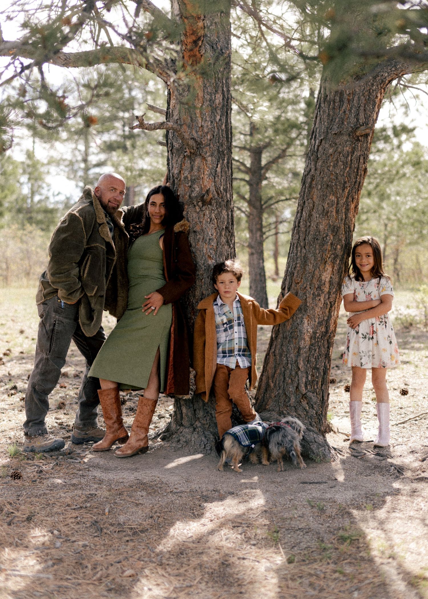 Family of four with a small dog posing by a large tree in a forest setting, wearing casual outdoor clothing.