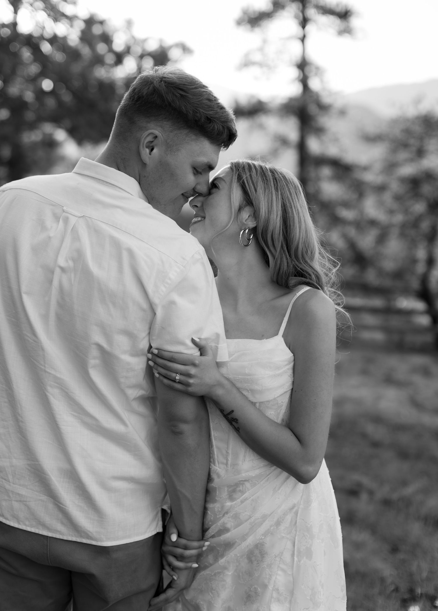 Black and white photo of a couple in romantic embrace, nearly kissing outdoors with trees and mountains in background.