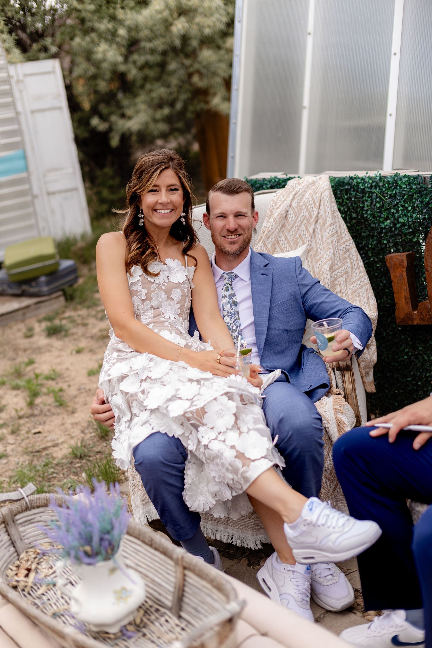 Smiling bride and groom in blue suit sit together holding champagne glasses at outdoor wedding reception.