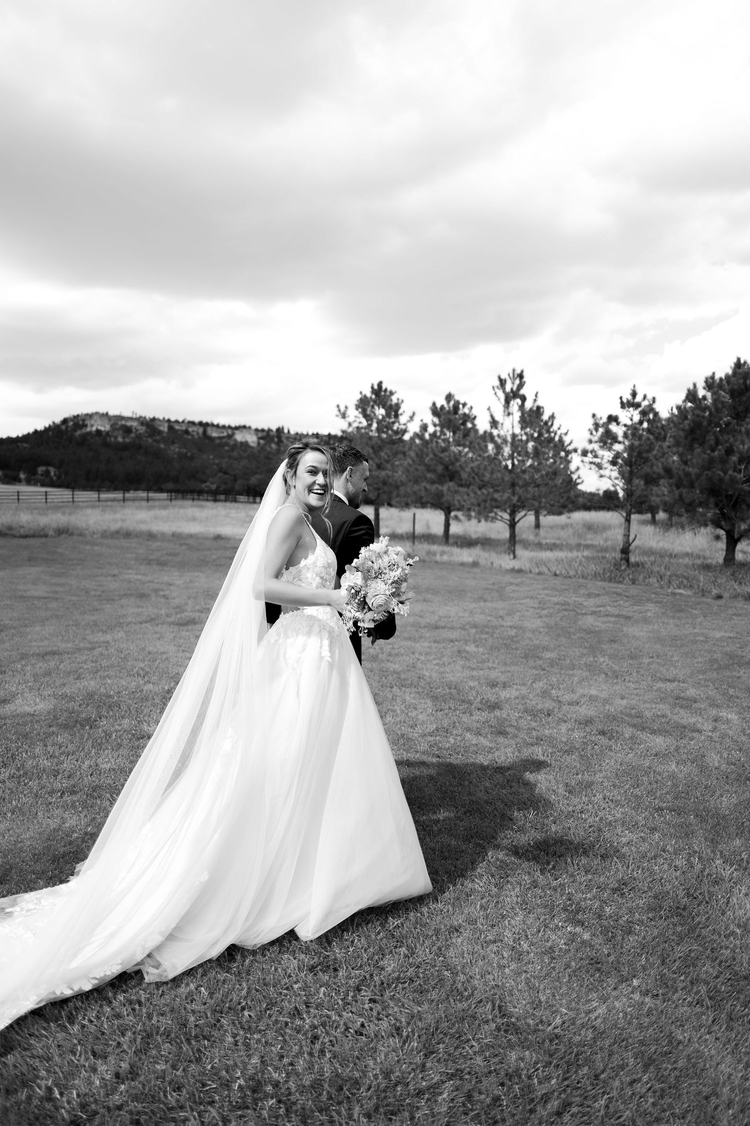 Bride in elegant white gown with veil smiles as groom embraces her in a lush outdoor setting.
