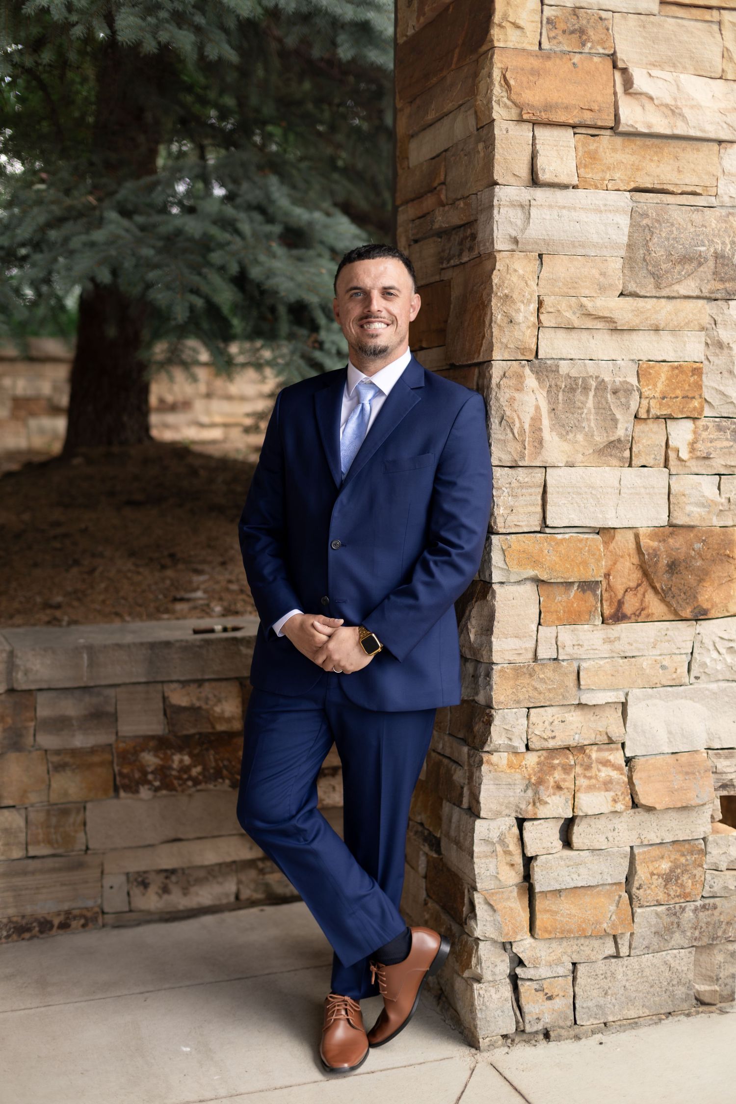 A man in a navy blue suit and brown shoes leans against a stone pillar, smiling outdoors.