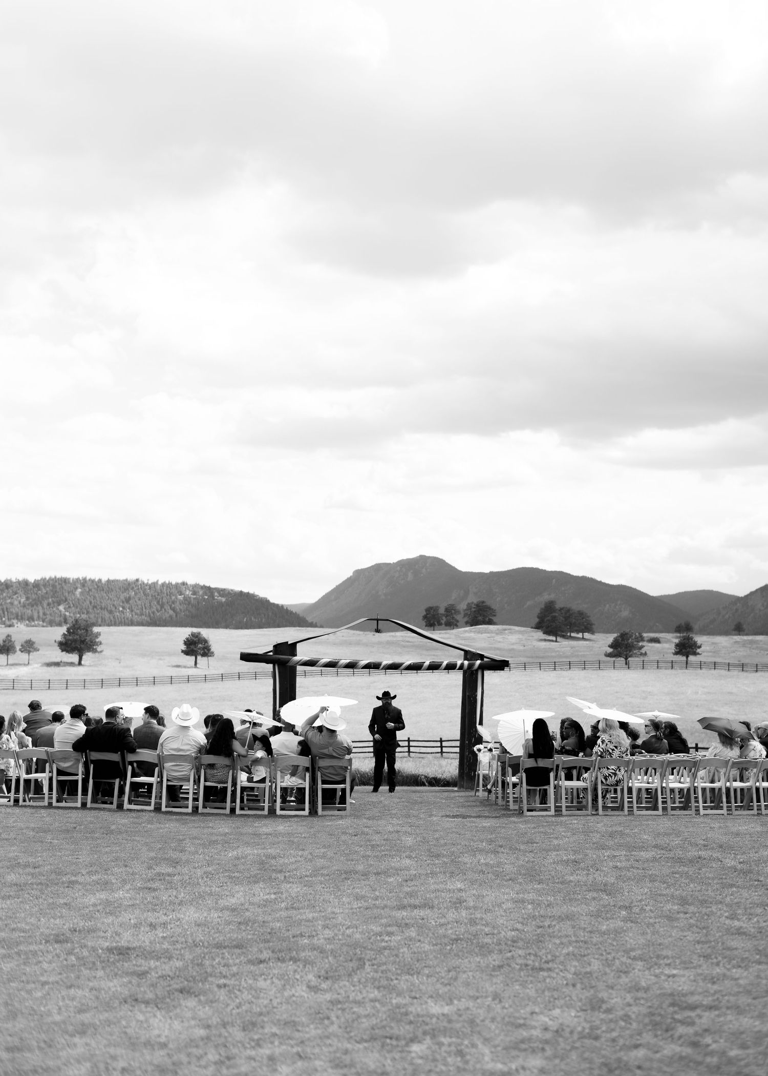 Black and white outdoor wedding ceremony on a ranch with mountain views, guests seated in rows under cloudy skies.
