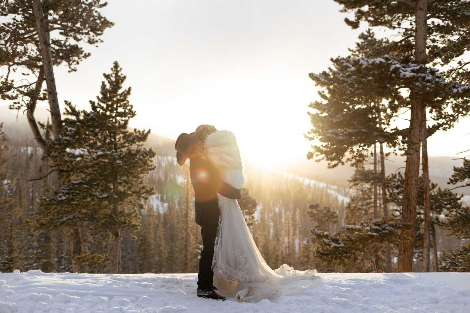 A couple embraces in a snowy winter forest landscape with golden sunlight streaming through evergreen trees.
