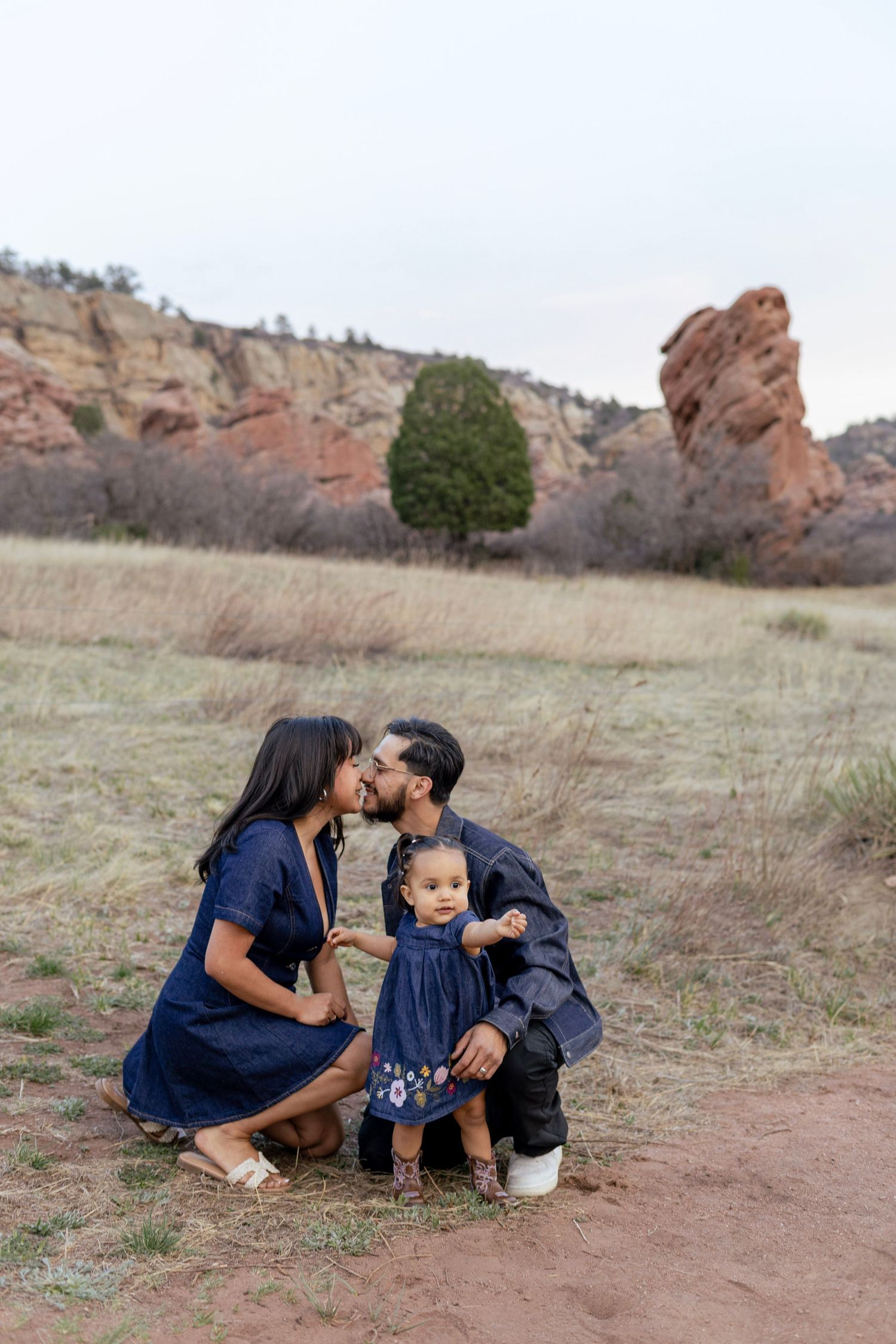 Family of three poses outdoors at Red Rocks, parents kissing while toddler stands between them in matching navy outfits.