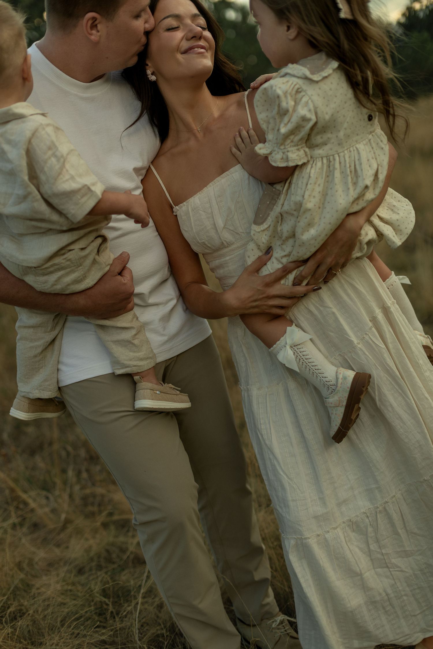 Happy family of four in neutral tones, parents holding two young children outdoors in golden light.