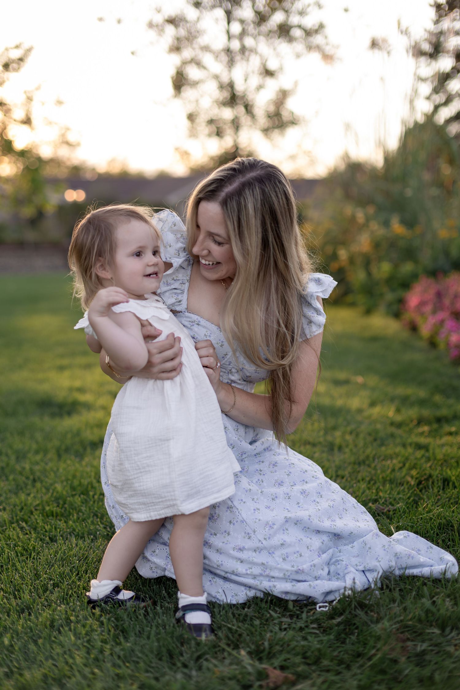 A smiling mother in a floral dress kneels on green grass, lovingly holding her toddler daughter in a white dress at sunset.