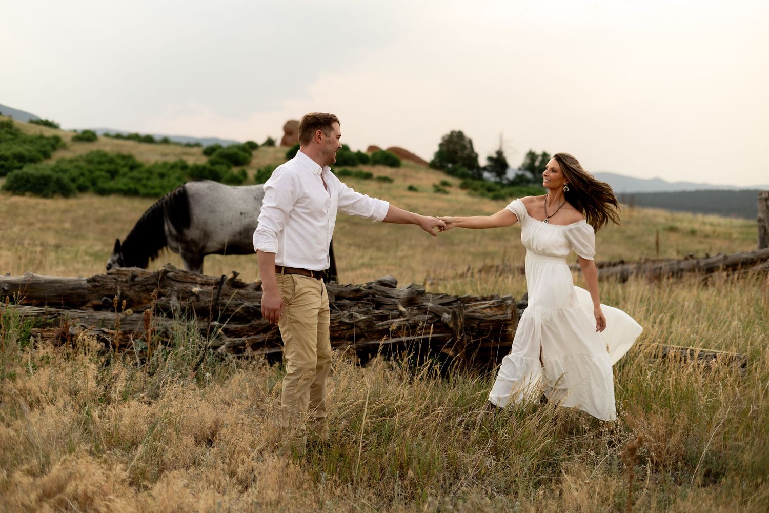 Couple holding hands dancing in a field with a horse in the background during a romantic outdoor engagement photo session.