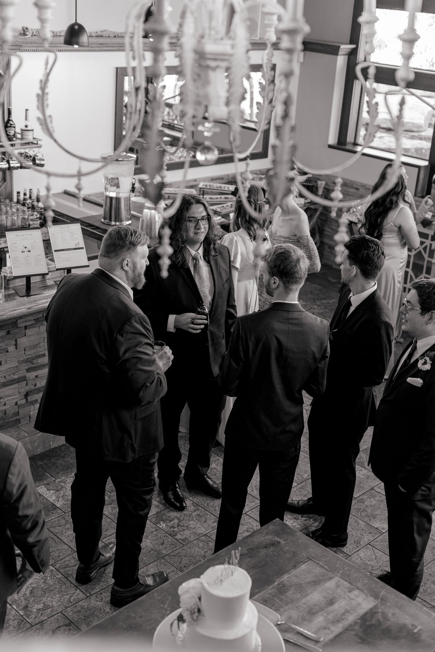 Black and white photo of groomsmen in suits conversing near wedding cake at reception venue.
