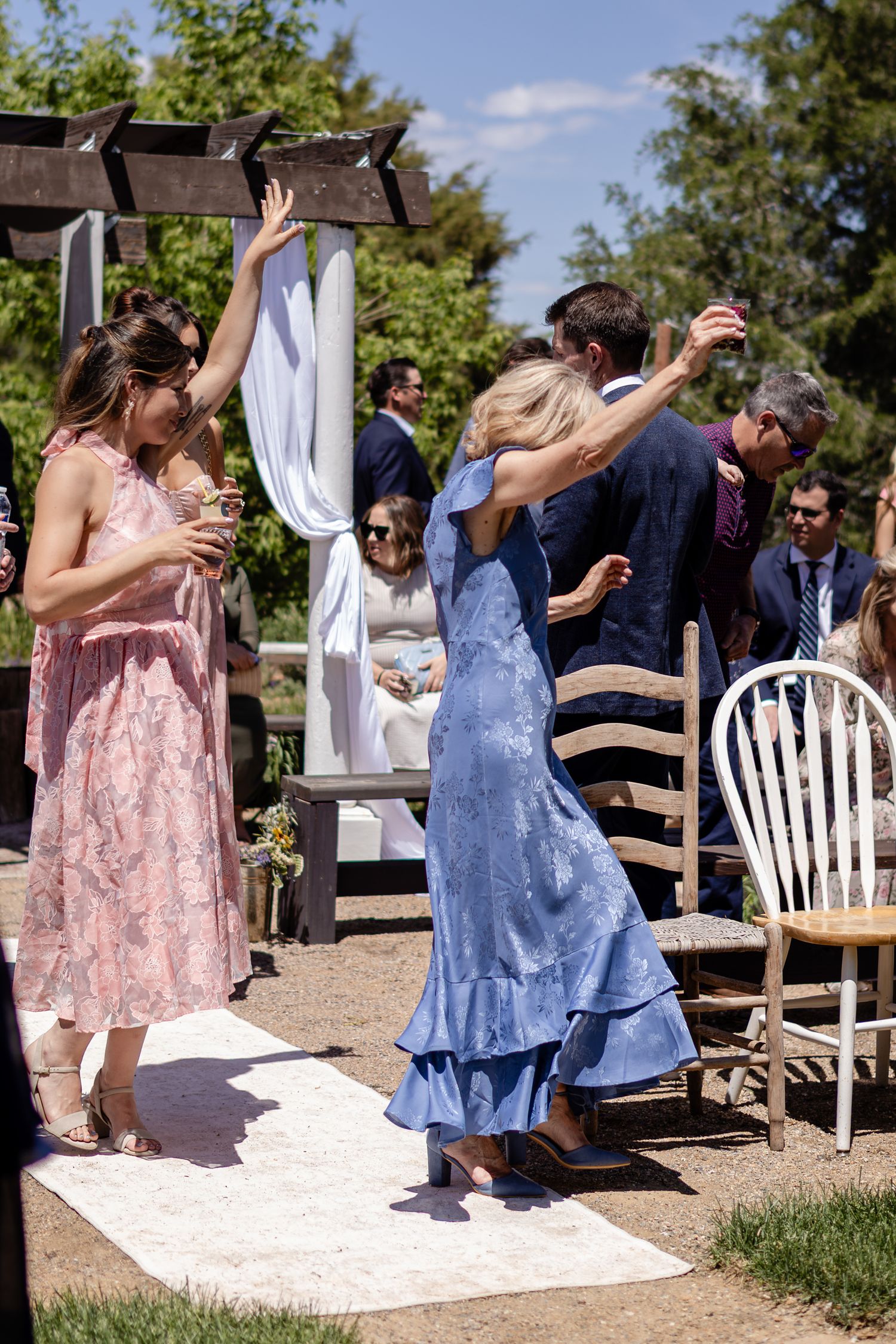 Guests dancing outdoors at a wedding ceremony, wearing formal attire near a wooden arbor on a sunny day.
