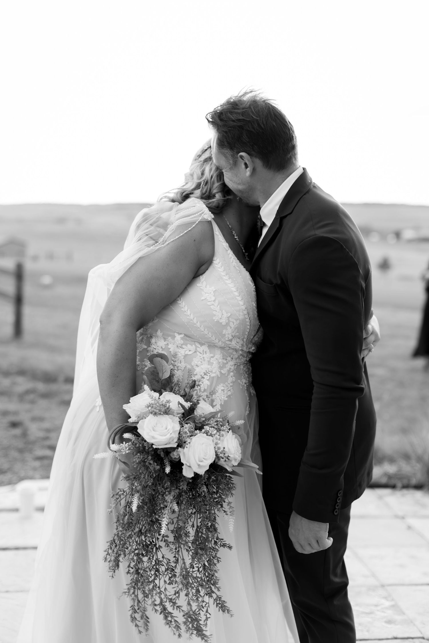 Black and white photo of a bride and groom sharing a kiss while she holds a cascading floral bouquet outdoors.