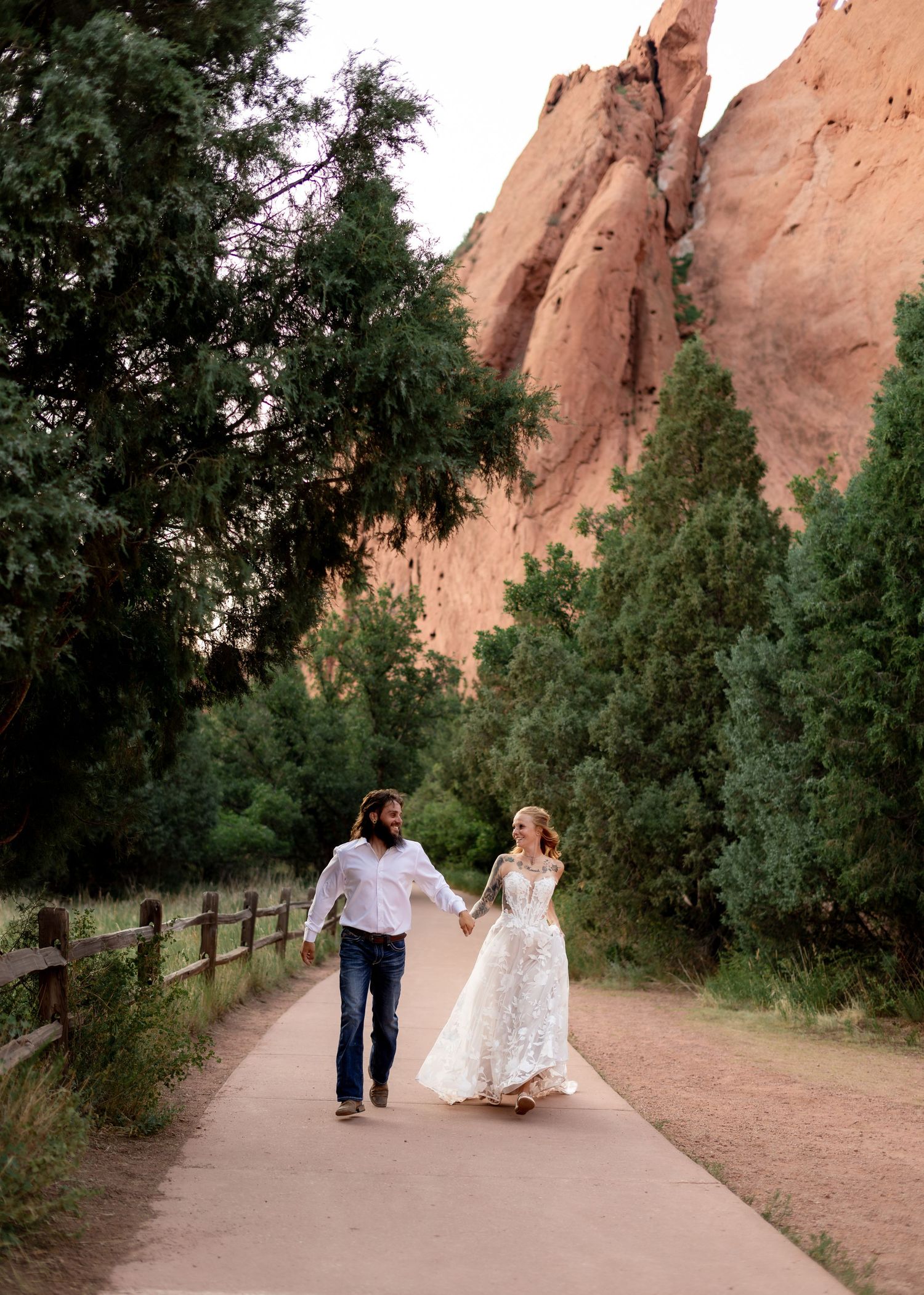 Couple in wedding attire holding hands walking on a path at Garden of the Gods with red rock formations and trees.