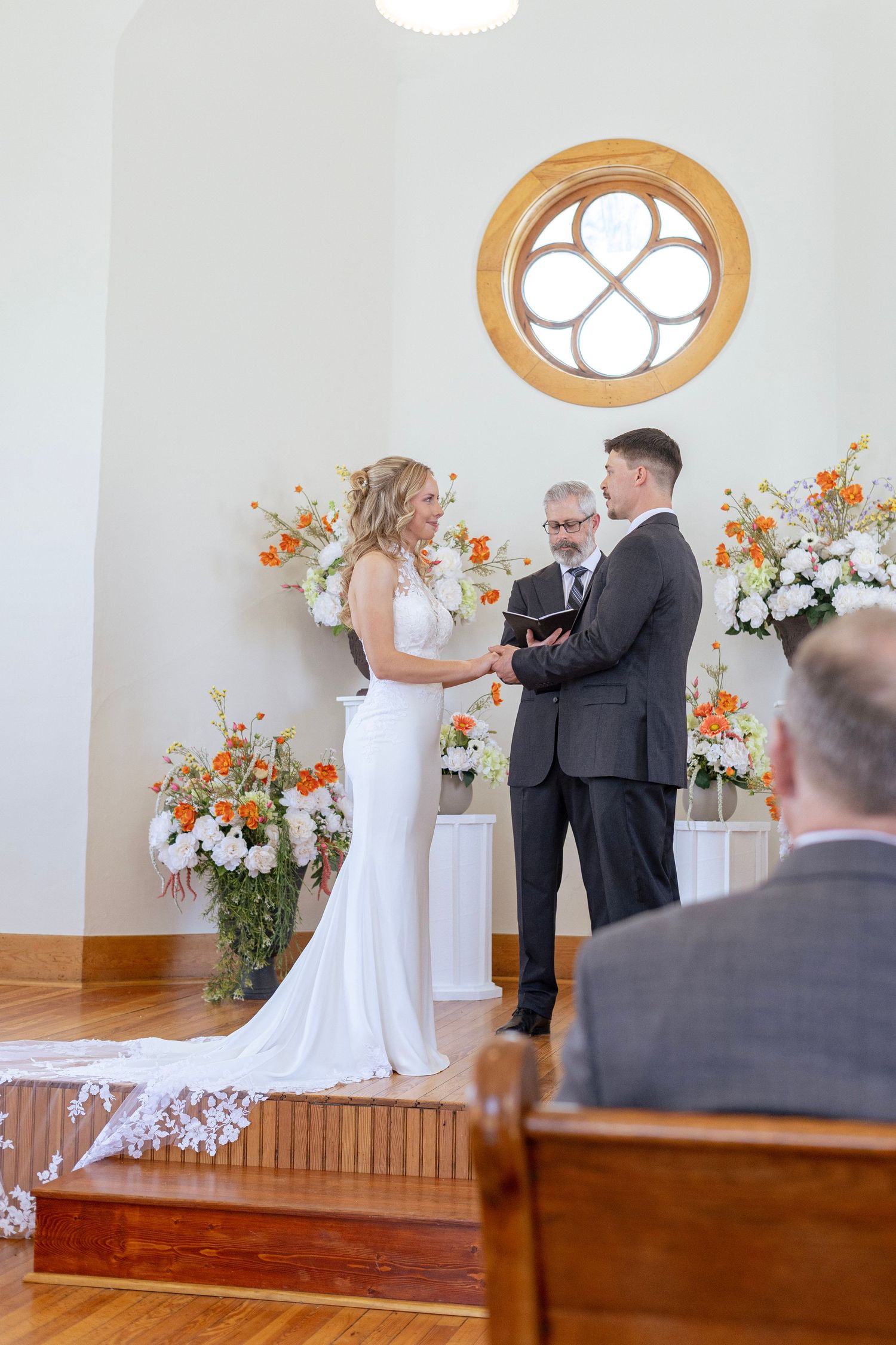 Bride and groom exchange vows at chapel altar with orange and white floral arrangements.