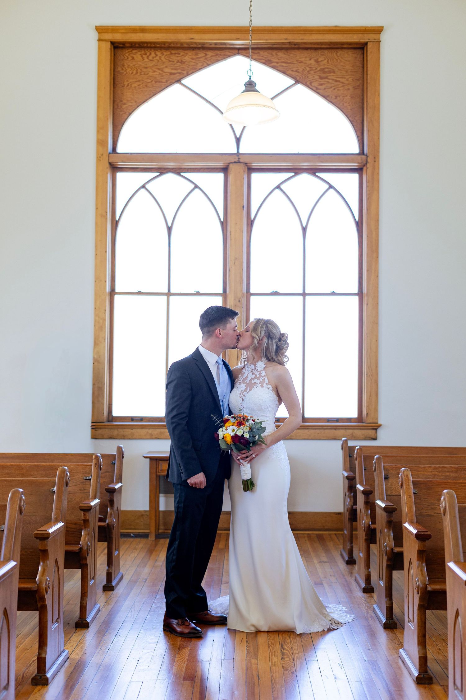 Bride and groom kissing in chapel aisle before large arched window with wooden pews.