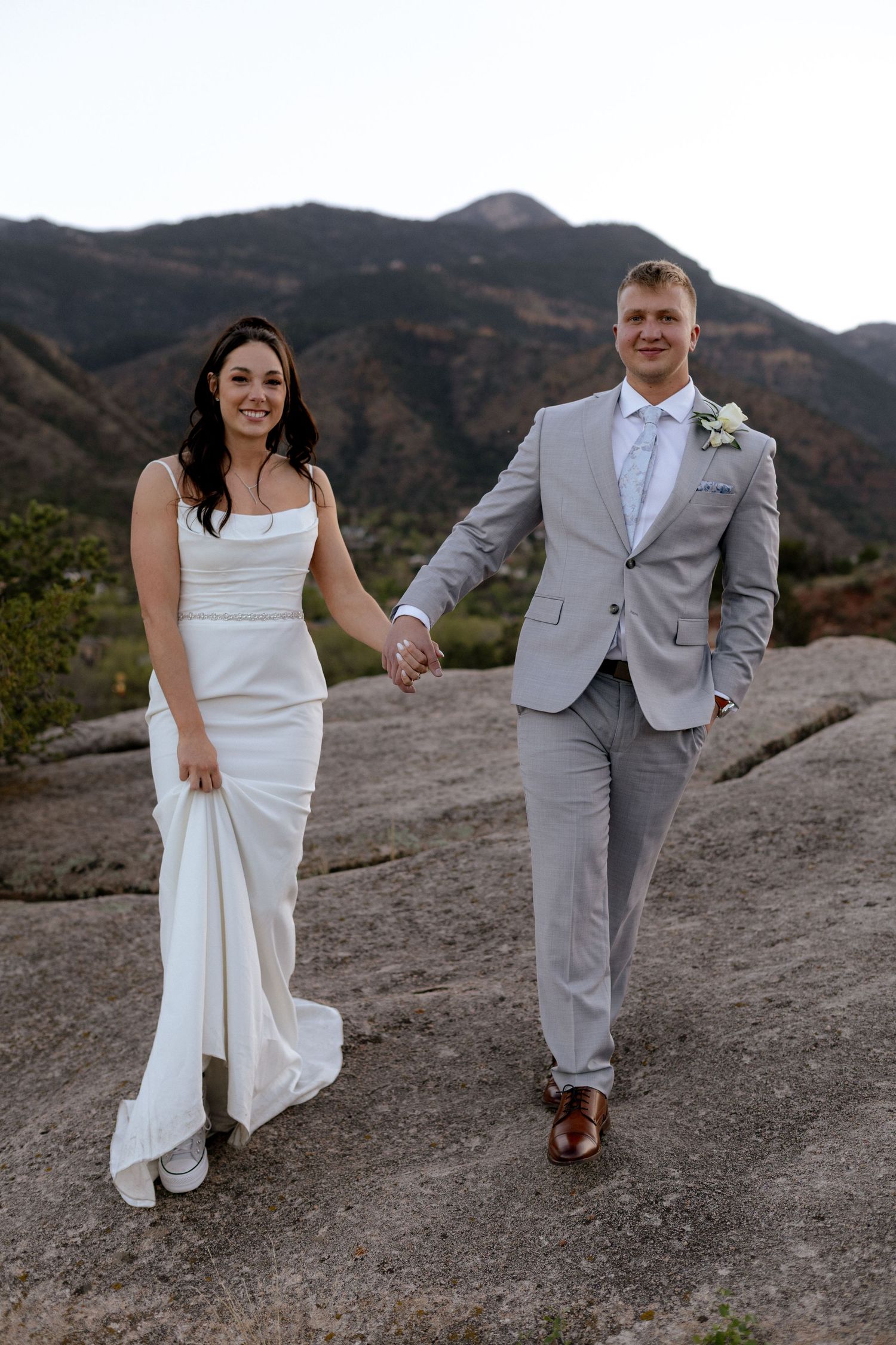 Bride and groom hold hands walking on rocky terrain with scenic mountain landscape behind them.