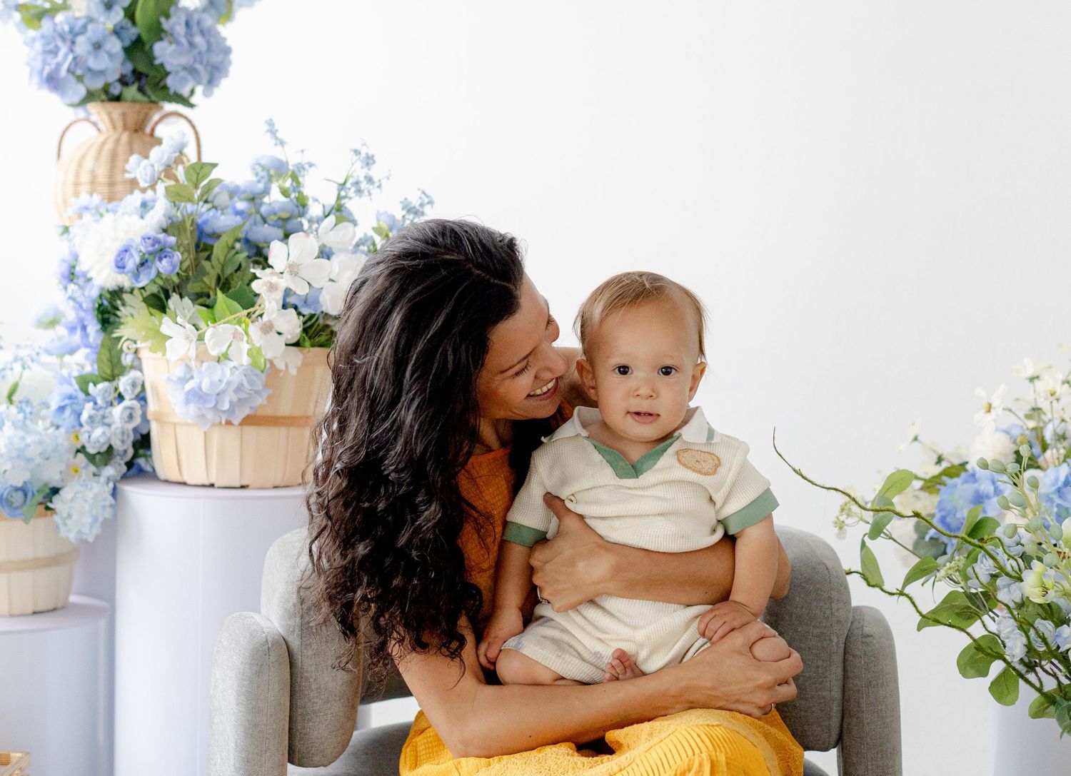 A mother kisses her baby boy surrounded by blue hydrangeas in a bright, elegant portrait session.