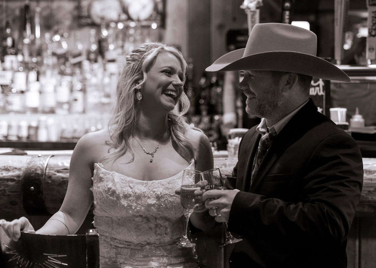 A bride and cowboy share a laugh at a bar counter in black and white photography.