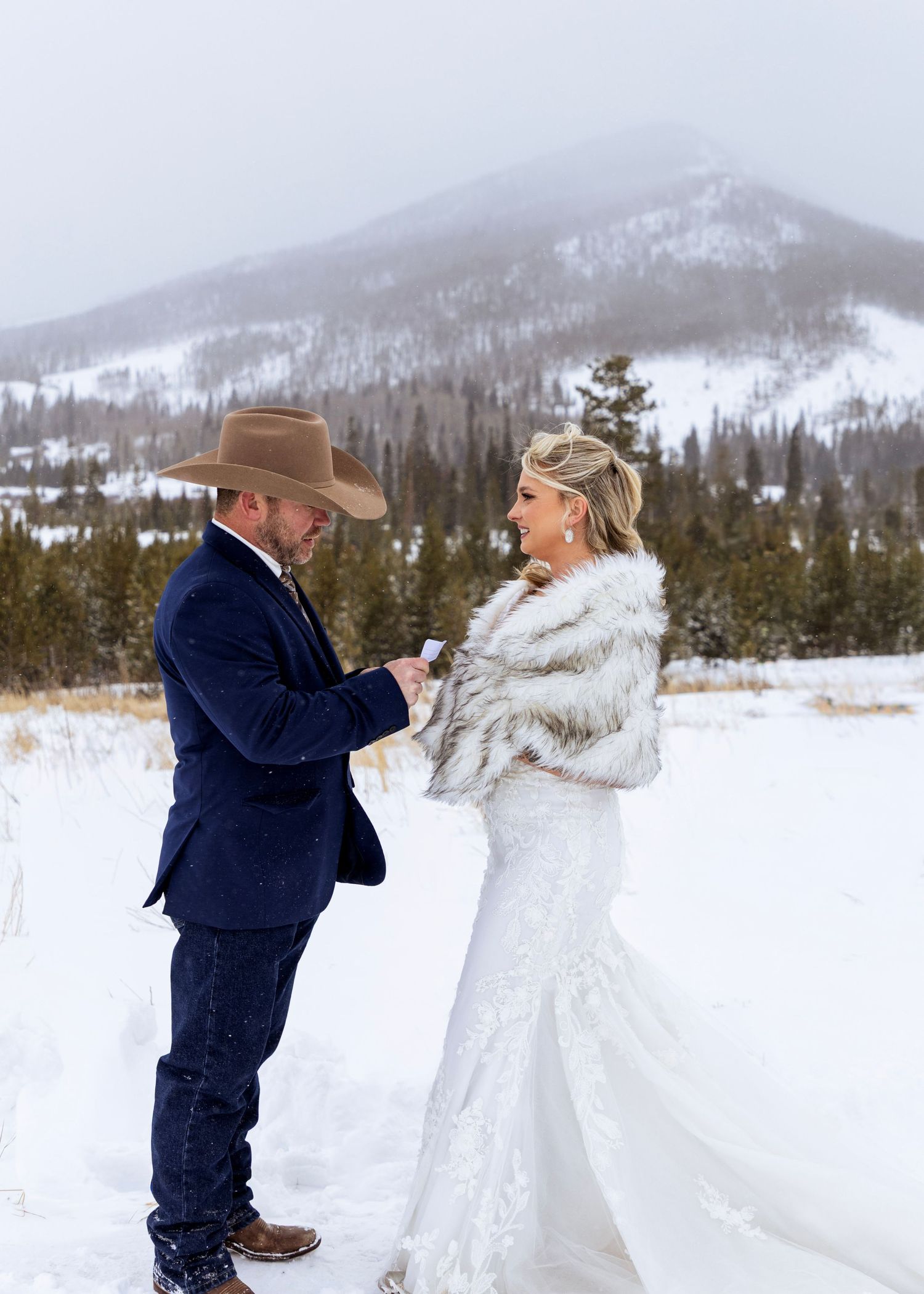 A winter wedding scene with a couple standing in snow with mountains and evergreen forest in the background.
