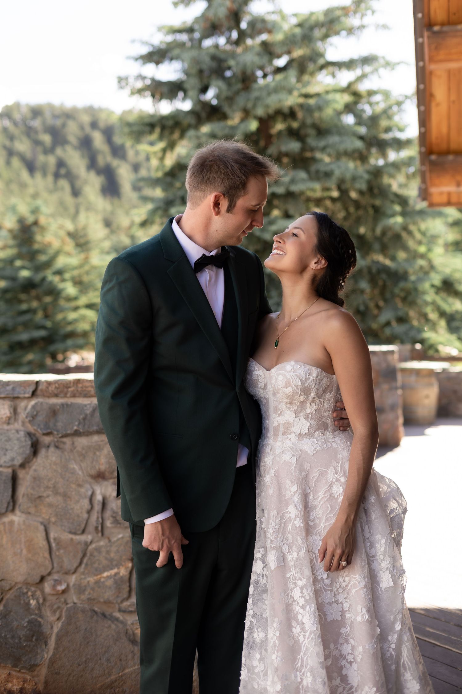 Bride and groom share a romantic moment outdoors, smiling at each other in elegant wedding attire with evergreen trees behind.
