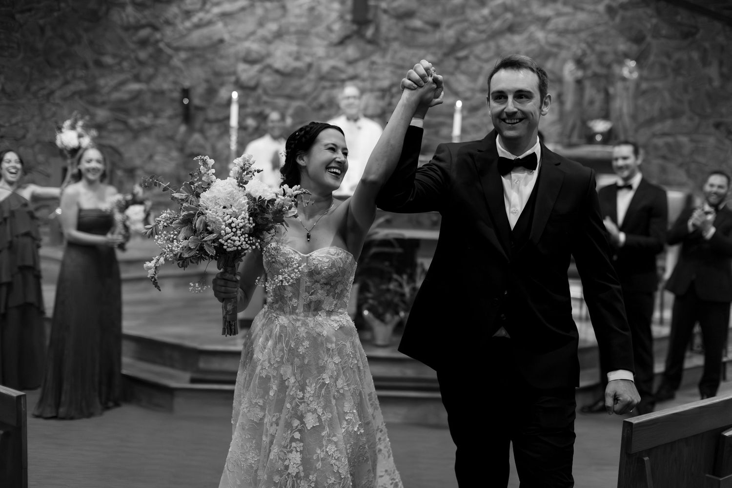 Joyful newlyweds celebrate at wedding ceremony exit, bride in lace gown holding bouquet, groom in tuxedo, black and white photo.