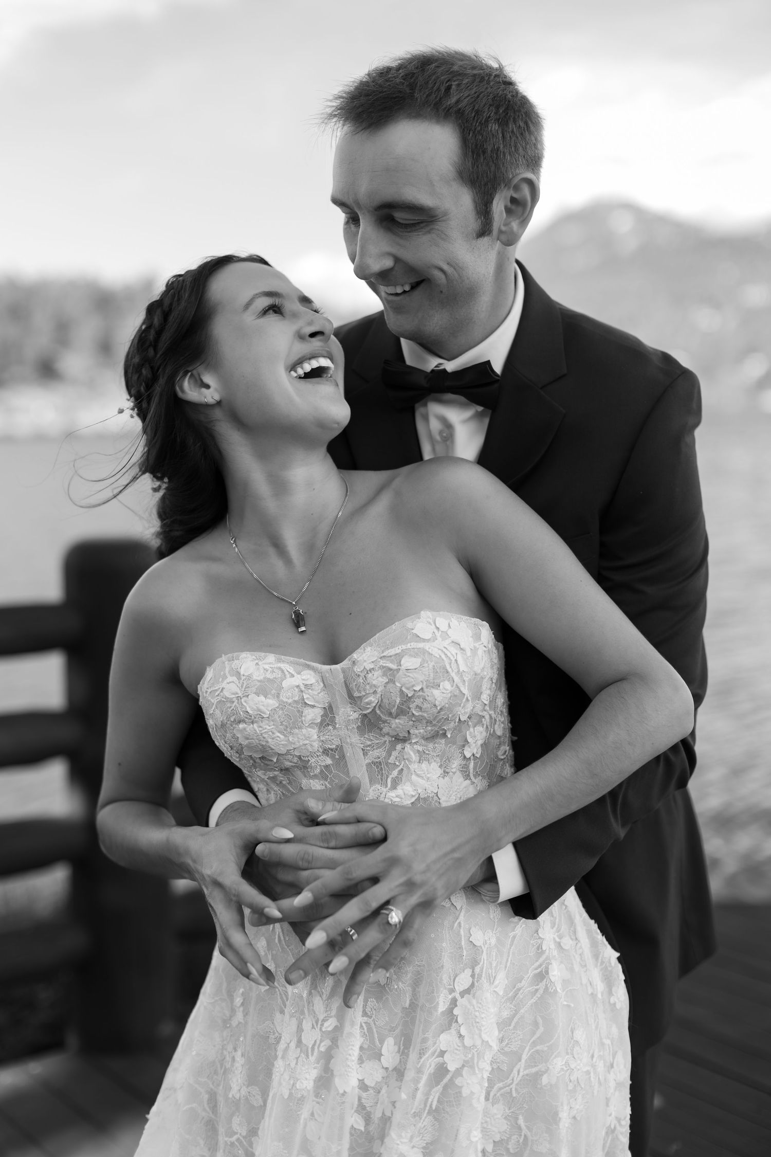 Black and white photo of a laughing bride in strapless lace gown embraced by groom in tuxedo on outdoor dock.